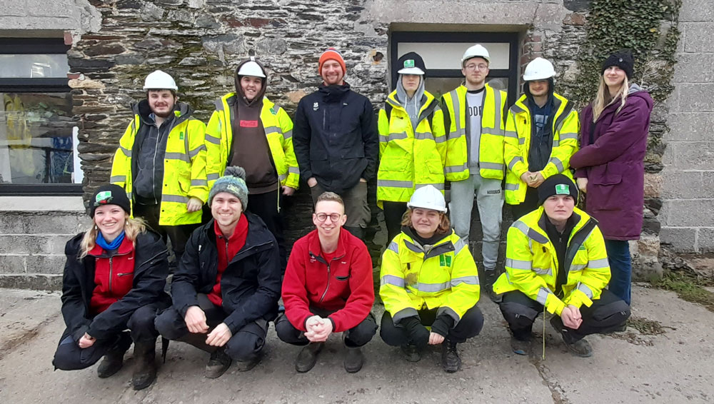 Group of 12 apprentices from the Plymouth Green Grid project, some with hard hats, standing against a wall. 