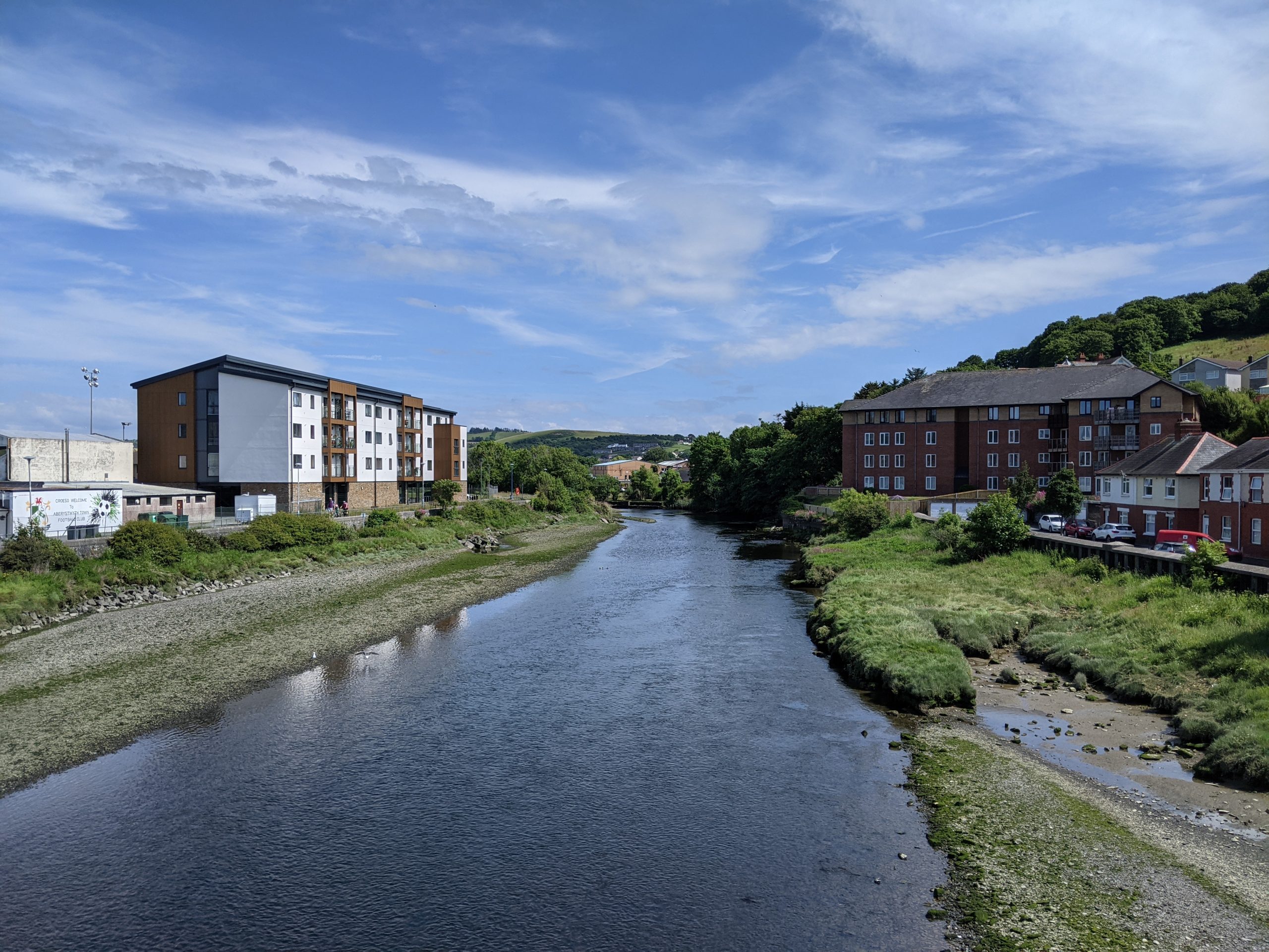 Wide river running through town with green space either side, modern buildings either side and a slightly cloudy blue sky.