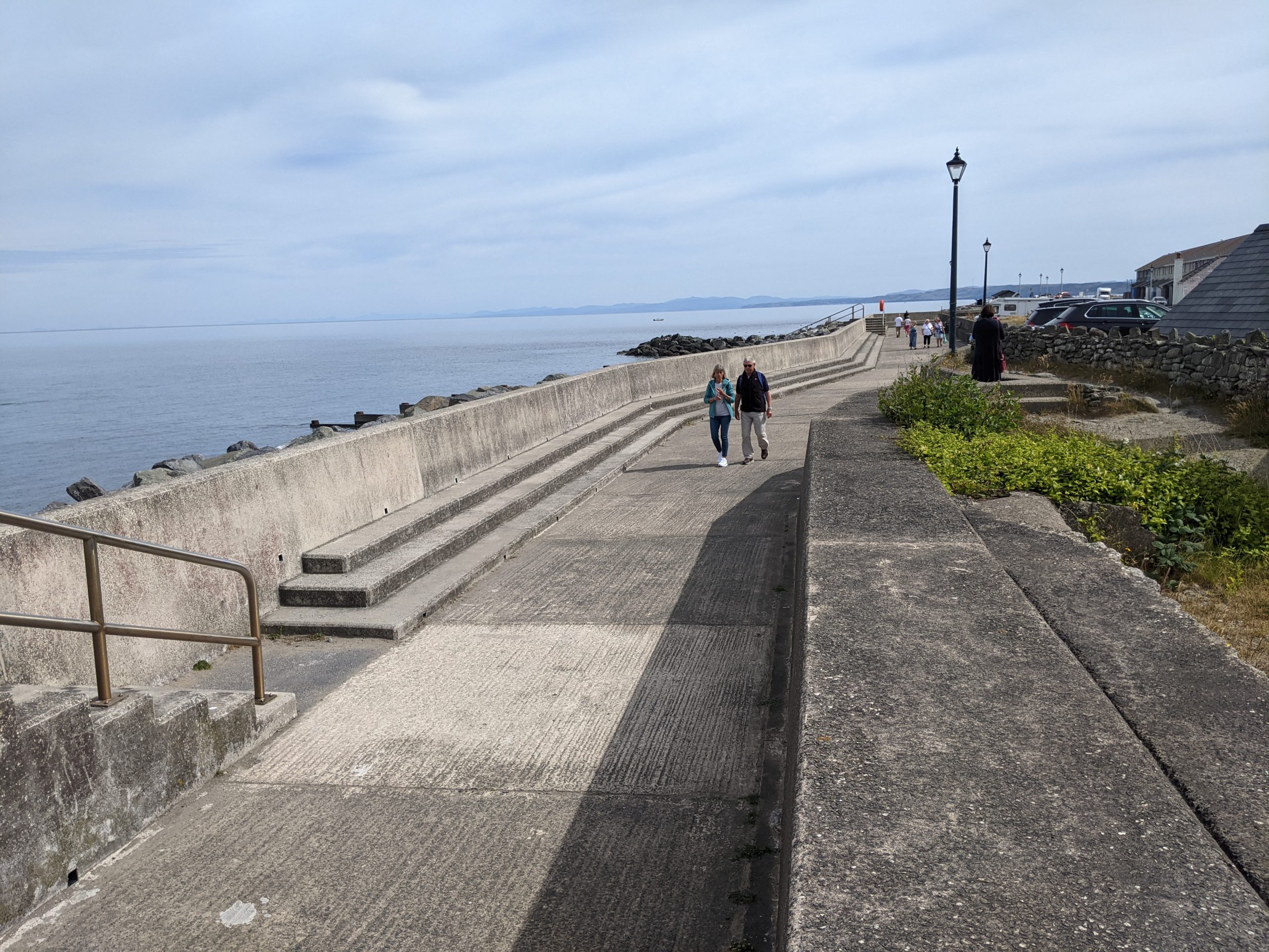 Concrete seafront promenade with lampposts, cars in the background and people walking along it.